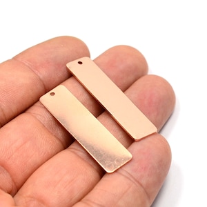 Puede incluir: Dos etiquetas rectangulares de metal de color oro rosa con un agujero en la parte superior. Las etiquetas están sobre una mano.