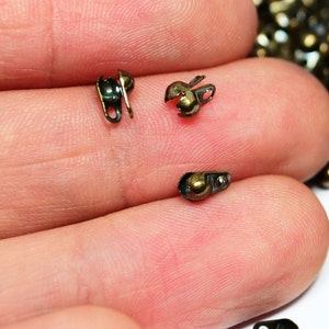 May include: Four antique gold-toned metal clasps with a rounded ball shape on one end and a loop on the other end. The clasps are on a finger.