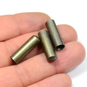 May include: Three small, cylindrical metal tubes with a bronze finish. The tubes are of varying lengths and are held in a person's hand against a white background. The tubes have a rounded end and a hollow end.
