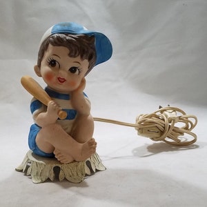 Könnte beinhalten: Eine Keramikfigur eines Jungen, der eine blaue Baseballkappe trägt und einen Holzschläger hält. Die Figur sitzt auf einem weißen Metallsockel und hat ein Kabel daran befestigt, was darauf hindeutet, dass es sich um ein Nachtlicht handelt.