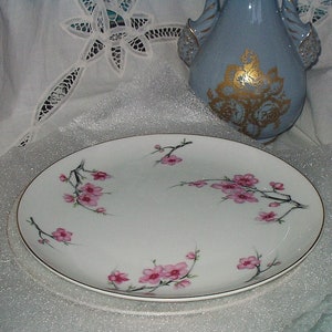 Puede incluir: Plato ovalado blanco para servir con borde dorado, decorado con ramas de flores de cerezo rosas. El plato tiene un delicado diseño floral, perfecto para servir aperitivos o postres. Un jarrón azul claro con detalles dorados está en el fondo.