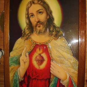 Puede incluir: Una imagen enmarcada de Jesucristo con un halo dorado, vistiendo una túnica roja con ribetes dorados y un corazón con llamas en el pecho. La imagen está sobre un fondo oscuro con un halo dorado alrededor de su cabeza.