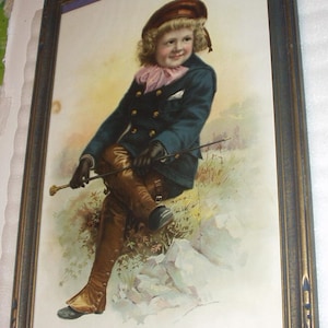 May include: A framed vintage print of a young child dressed in a blue jacket and brown riding pants, holding a riding crop. The child is sitting on a rock with a background of green foliage. The print is titled "The Young Cavalier".