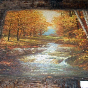 May include: A framed painting depicting a woodland scene with a stream flowing through it. The trees are in autumnal colours, with shades of orange, yellow, and red. The stream is clear and flowing, with a few rocks in the water.