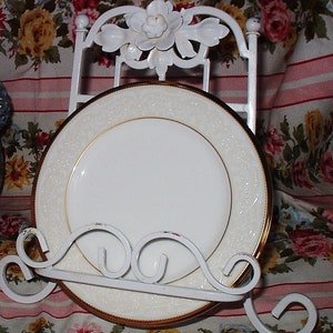 May include: White ceramic plate with a gold rim and intricate design, displayed on a white metal plate stand with a decorative floral design.