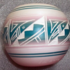 May include: A small, round, pink and white ceramic pot with a teal design of stylized fish. The pot has a smooth, glossy finish.