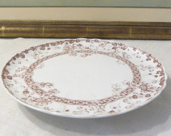 May include: A round white ceramic plate featuring a brown floral transferware pattern along the scalloped rim and a central circular border. The design includes intricate botanical details and lace-like motifs printed in a monochromatic brown ink.