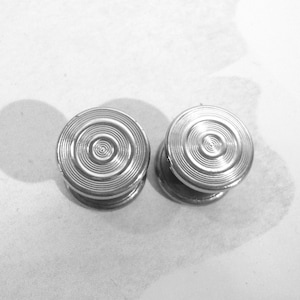 May include: Two circular silver-toned metal buttons featuring a concentric ring pattern on the top surface. Each button has a smooth, raised center and a series of fine, etched circular grooves radiating outward toward the beveled edge.