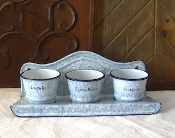 May include: A vintage blue and white enamelware caddy with three small pots. Each pot has a blue rim and text in French. The caddy is on a white surface with red stripes, with a wooden background.