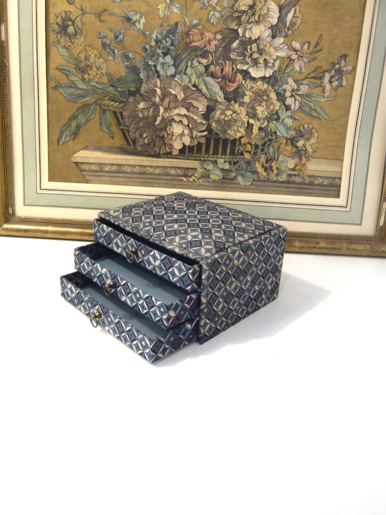 May include: A small, blue jewelry box with two pull-out drawers. The box and drawers are covered in a geometric diamond pattern. The box has small, gold-colored drawer pulls. A framed floral print painting is in the background.