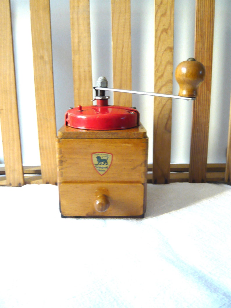 May include: A vintage wooden coffee grinder with a red top and a silver handle. The grinder has a drawer and a wooden knob. The Peugeot logo is visible on the front. The grinder is made of wood and metal.