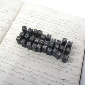 May include: A selection of vintage metal letterpress printing blocks, displayed on an open book with handwritten script. The blocks are black with white lettering, including numbers and letters. The book's pages are aged and yellowed.