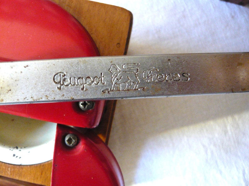 May include: A vintage Peugeot Freres coffee mill featuring a bright red metal top and a wooden base. A horizontal silver metal handle is engraved with the text Peugeot Freres and a lion logo. The mechanism includes visible screws.