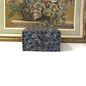 May include: A small, two-drawer jewelry box with a geometric pattern in shades of blue, green, and pink. The box has brass-colored drawer pulls. A framed floral print is in the background.