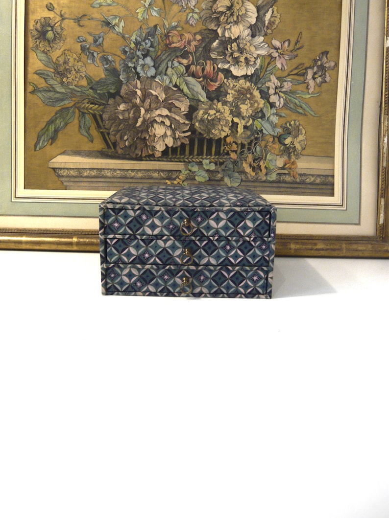 May include: A small, two-drawer jewelry box with a geometric pattern in shades of blue, green, and pink. The box has brass-colored drawer pulls. A framed floral print is in the background.
