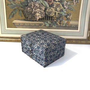 May include: A rectangular box with a geometric pattern in shades of blue and gray. The box has a hinged lid and small metal clasps. A framed floral print with a gold frame is in the background.