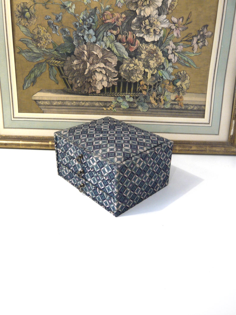May include: A rectangular box with a geometric pattern in shades of blue and gray. The box has a hinged lid and small metal clasps. A framed floral print with a gold frame is in the background.