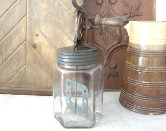 Könnte beinhalten: Eine antike Buttermaschine aus Glas mit einem Metalldeckel und Zahnrädern. Das transparente Glasgefäß hat eine sechseckige Form und einen Metallrührer im Inneren. Die Buttermaschine steht vor einem hölzernen Hintergrund mit dekorativen Paneelen.