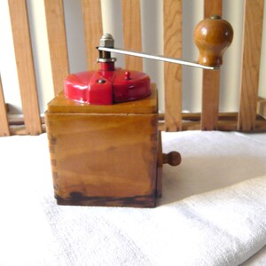 May include: A vintage wooden coffee grinder with a red top and a wooden handle. The grinder has a rectangular wooden base and a metal crank arm. The wooden handle is a rounded shape. The grinder is sitting on a white cloth.