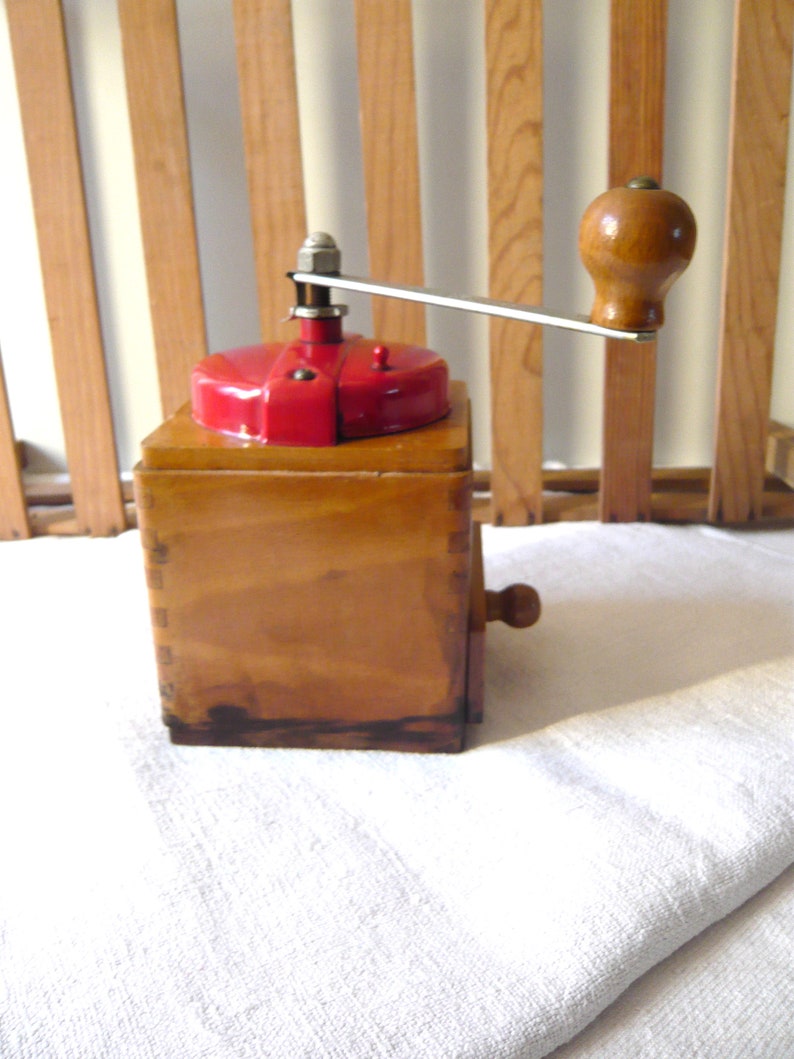 May include: A vintage wooden coffee grinder with a red top and a wooden handle. The grinder has a rectangular wooden base and a metal crank arm. The wooden handle is a rounded shape. The grinder is sitting on a white cloth.