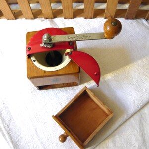 May include: A wooden coffee grinder with a square base, a red metal top lid, and a silver metal crank handle featuring the engraved text Peugeot Freres. The device includes a removable wooden drawer and a white interior grinding chamber.