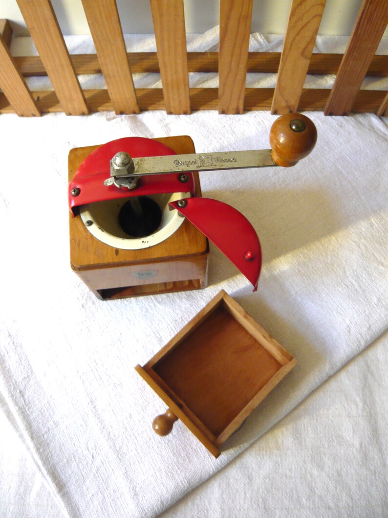 May include: A wooden coffee grinder with a square base, a red metal top lid, and a silver metal crank handle featuring the engraved text Peugeot Freres. The device includes a removable wooden drawer and a white interior grinding chamber.