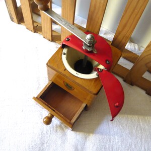 May include: A wooden coffee grinder featuring a light brown square base with a pull-out drawer, a white circular hopper, and a red metal lid. A silver metal crank handle with a round wooden knob is attached to the top, and a small lion logo is visible on the front.
