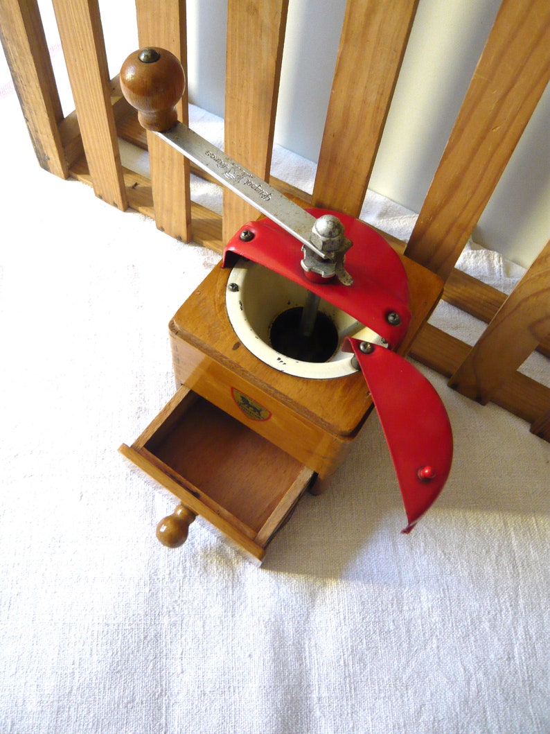 May include: A wooden coffee grinder featuring a light brown square base with a pull-out drawer, a white circular hopper, and a red metal lid. A silver metal crank handle with a round wooden knob is attached to the top, and a small lion logo is visible on the front.