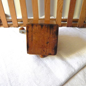 May include: A vintage wooden coffee grinder with a crank handle. The grinder is a warm brown color, showing signs of age and use. It has a rectangular body and a small, decorative foot.