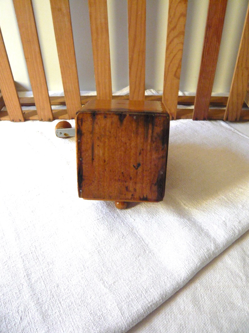 May include: A vintage wooden coffee grinder with a crank handle. The grinder is a warm brown color, showing signs of age and use. It has a rectangular body and a small, decorative foot.