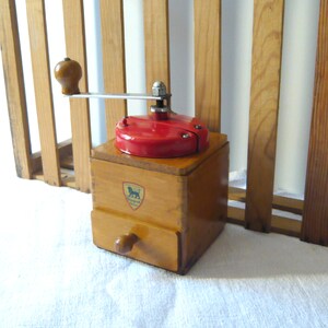 May include: A vintage wooden coffee grinder with a red top and a drawer. The grinder has a metal handle and a wooden knob. A crest with a lion is visible on the front. The grinder is made of light brown wood.