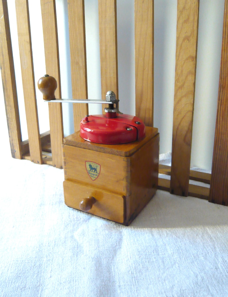 May include: A vintage wooden coffee grinder with a red top and a drawer. The grinder has a metal handle and a wooden knob. A crest with a lion is visible on the front. The grinder is made of light brown wood.