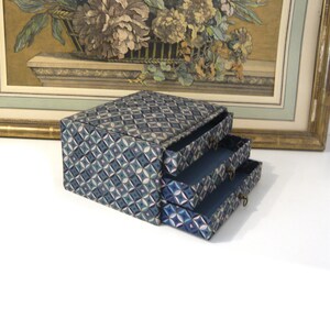 May include: A three-drawer jewelry box with a geometric pattern in shades of blue, teal, and white. The drawers are open, revealing a dark blue interior. The box sits in front of a framed floral print.