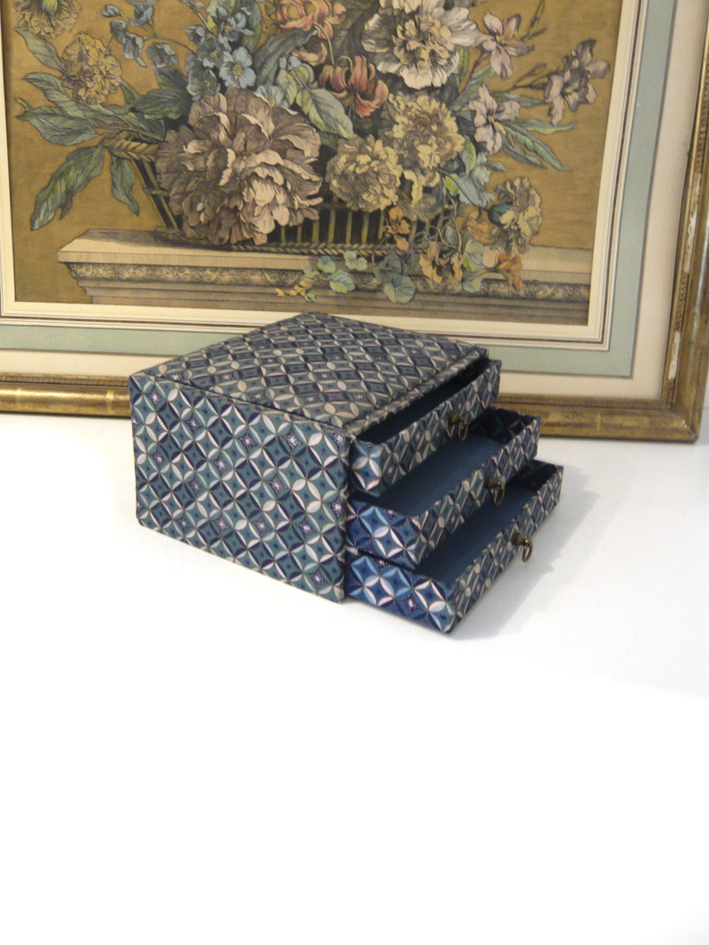 May include: A three-drawer jewelry box with a geometric pattern in shades of blue, teal, and white. The drawers are open, revealing a dark blue interior. The box sits in front of a framed floral print.