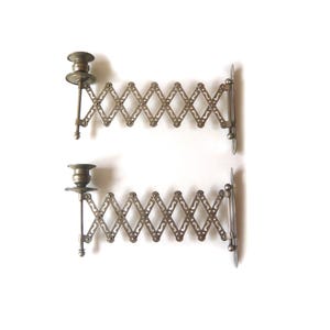 May include: A pair of wall-mounted candle holders made of metallic silver-toned material. Each holder features an expandable accordion-style arm with a lattice pattern and a circular base plate for attachment, topped with a small candle cup.
