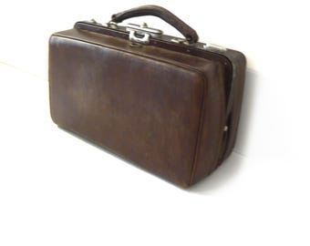 May include: A rectangular dark brown leather doctor bag featuring a structured frame, a single top handle, and a silver-toned metal latch closure mechanism. The bag has a smooth, solid-colored exterior surface with visible stitching along the edges.
