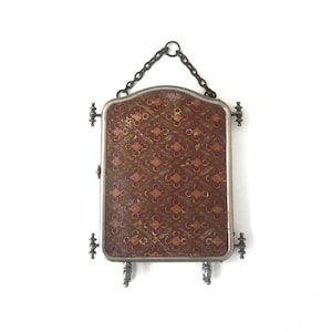 May include: An antique, rectangular mirror with a decorative, patterned fabric backing in shades of brown, red, and gold. The mirror is framed in silver metal and hangs from a chain. Decorative metal accents are on the sides and bottom.