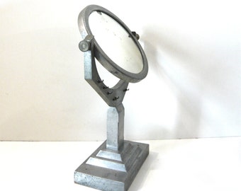 May include: A silver-colored, Art Deco-style vanity mirror. The round mirror is set in a thick frame and is supported by a rectangular base. The mirror has a pivoting design and small hooks on the frame.