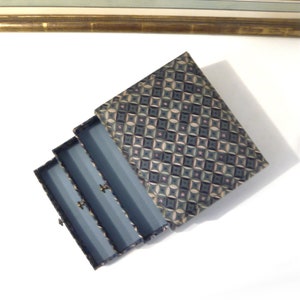 May include: An open jewelry box with a patterned lid. The box is blue with three compartments, each with a small gold-colored clasp. The lid features a geometric design in shades of blue, green, and beige.
