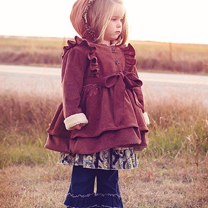 May include: A young girl wearing a brown corduroy jacket with a ruffled trim and a matching hood. The jacket has a brown ribbon tied at the waist. She is also wearing a blue and white patterned skirt and blue denim pants with ruffles at the bottom. The girl is standing in a field of tall grass.