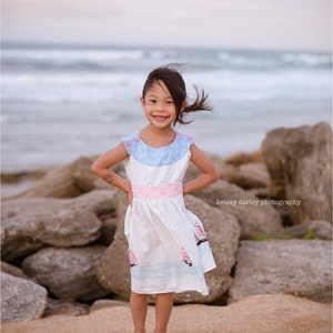 Puede incluir: Una niña pequeña con un vestido blanco con detalles azules y rosas y un estampado de velero está de pie en una playa rocosa. Su cabello está volando en el viento y ella está sonriendo a la cámara.