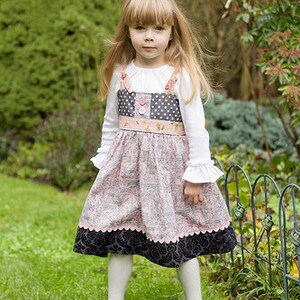 May include: A girl in a pink and grey patterned dress with a white long-sleeved top. The dress features a black scalloped hem, worn with white tights and black Mary Jane shoes. The image includes the text "The Queen Bee Photography".