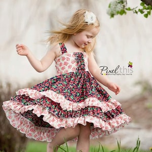 Könnte beinhalten: Ein junges Mädchen trägt ein mehrfarbiges Kleid mit Blumenmuster und Rüschen. Das Kleid hat eine dunkelblaue Basis mit roten und weißen Akzenten. Das Mädchen steht auf einer grasbewachsenen Wiese mit einem Baum im Hintergrund.