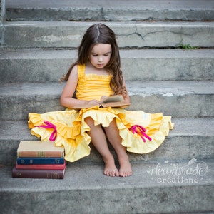 May include: A young girl in a yellow dress with pink accents sits on a set of concrete steps, reading a book. She is wearing a yellow dress with pink ruffles and a book in her hands. The book is open and she is looking at it. There are three books stacked on the steps below her.