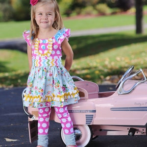 May include: A young girl wearing a pink, blue, and yellow dress with a white polka dot top and white polka dot leggings stands next to a pink toy car. The car has a silver steering wheel and the words "Bearing Drive" and "Pretty in Pink" on the side.