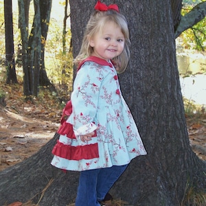 Pode incluir: Uma menina pequena vestindo um vestido estampado em azul e vermelho com um laço vermelho no cabelo. Ela está em pé na frente de uma árvore e sorrindo para a câmera.
