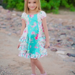 May include: A young girl wearing a light blue and white floral dress with pink ruffles and a pink bow in her hair. She is standing on a gravel path and smiling at the camera.