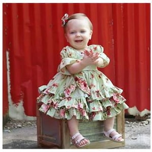 May include: A little girl wearing a green floral dress with a ruffled skirt and a bow in her hair. She is sitting on a wooden box in front of a red wall.