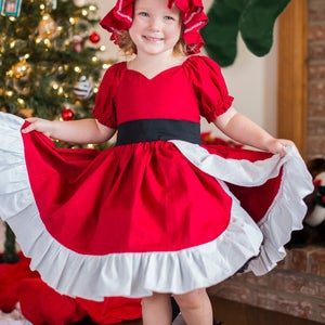 May include: A young girl wearing a red dress with a white ruffle trim and a red bonnet. She is standing in front of a Christmas tree and holding the skirt of her dress.