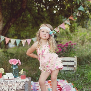 May include: A young girl wearing a pink floral dress with a white pearl necklace and a blue and white flower headband stands on a colorful patchwork quilt in a grassy area.